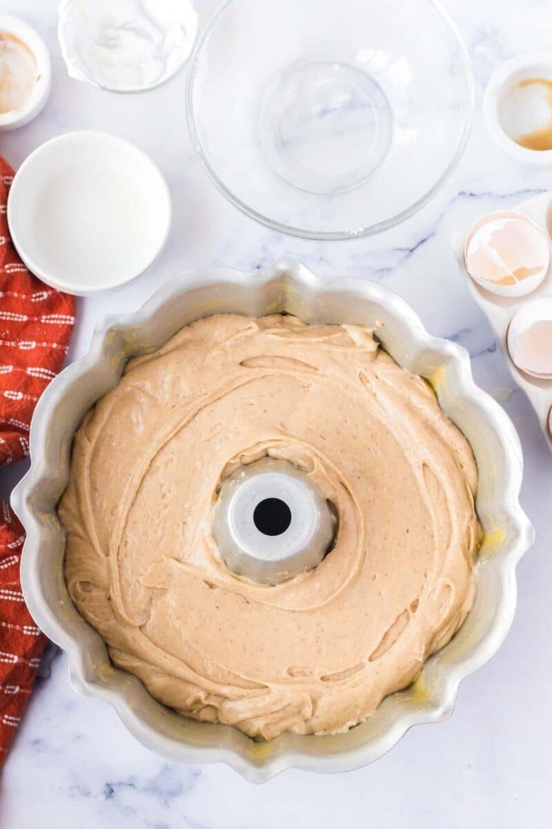 Bundt pan filled with unbaked cake batter on a marble countertop, surrounded by baking ingredients, a mixing bowl, cracked eggshells, and a red dish towel.