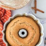 A baked bundt cake in a scalloped pan sits on a marble surface near a red towel, a wooden tray, a plate with cinnamon sticks, and forks.