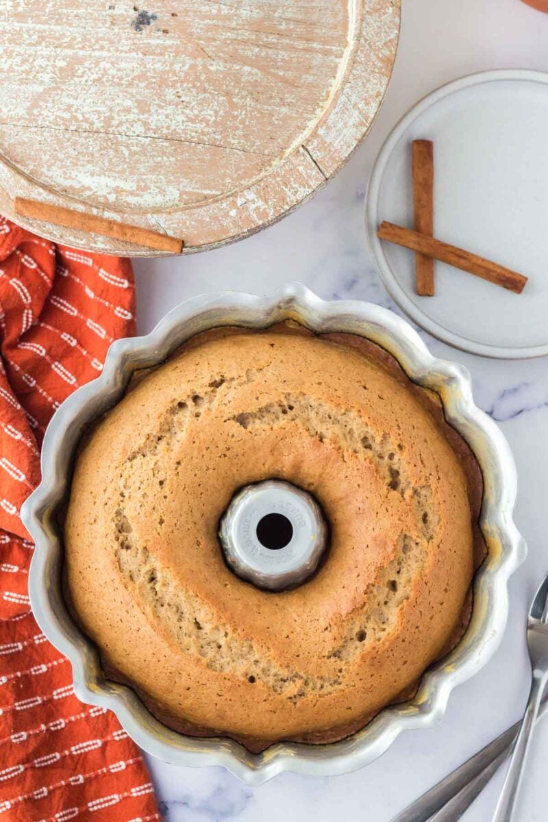 A baked bundt cake in a scalloped pan sits on a marble surface near a red towel, a wooden tray, a plate with cinnamon sticks, and forks.