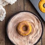 A baked donut sits on a plate of cinnamon sugar, ready to be coated, with a donut pan and a cloth napkin nearby on a wooden surface.