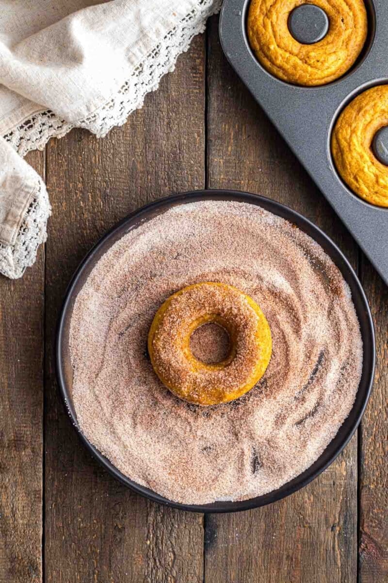 A baked donut sits on a plate of cinnamon sugar, ready to be coated, with a donut pan and a cloth napkin nearby on a wooden surface.