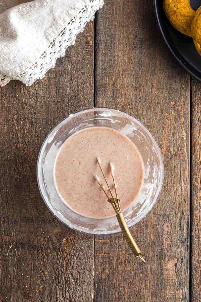 A glass bowl of brown batter with a small whisk, on a rustic wooden surface next to a beige cloth and a black plate.