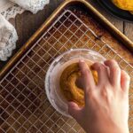 A hand dips a round pumpkin donut, into a bowl with a baking tray and wire rack underneath. Other similar items are on a black plate nearby.