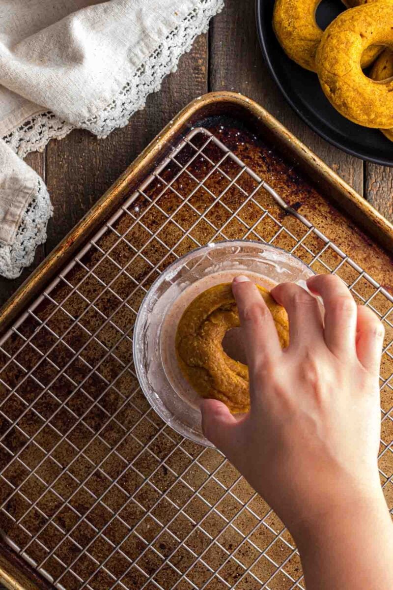 A hand dips a round pumpkin donut, into a bowl with a baking tray and wire rack underneath. Other similar items are on a black plate nearby.