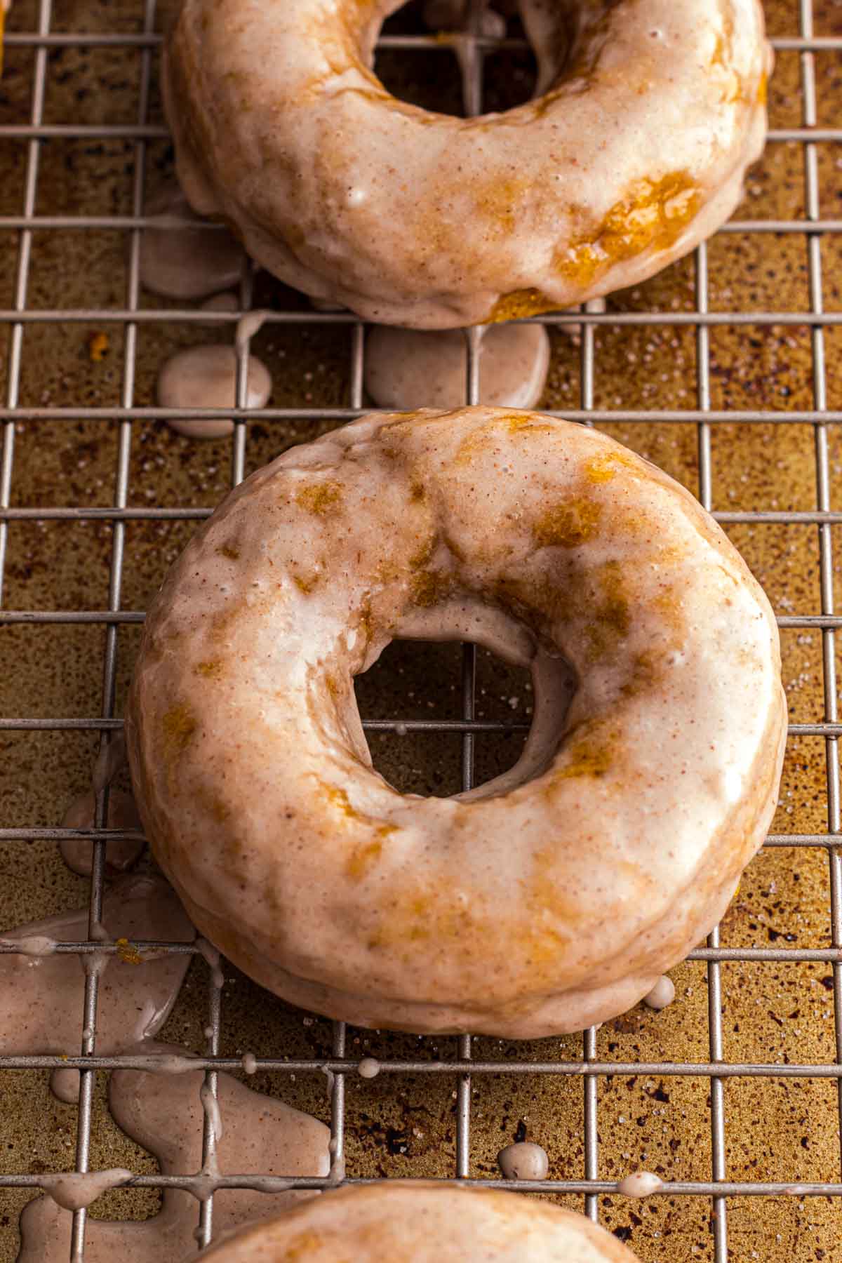 Two glazed pumpkin donuts rest on a wire cooling rack with glaze drips visible on the rack and surface below.