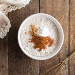 A white bowl with flour, baking powder, cinnamon, and nutmeg on a wooden surface, next to a metal whisk and a cloth napkin with lace trim.