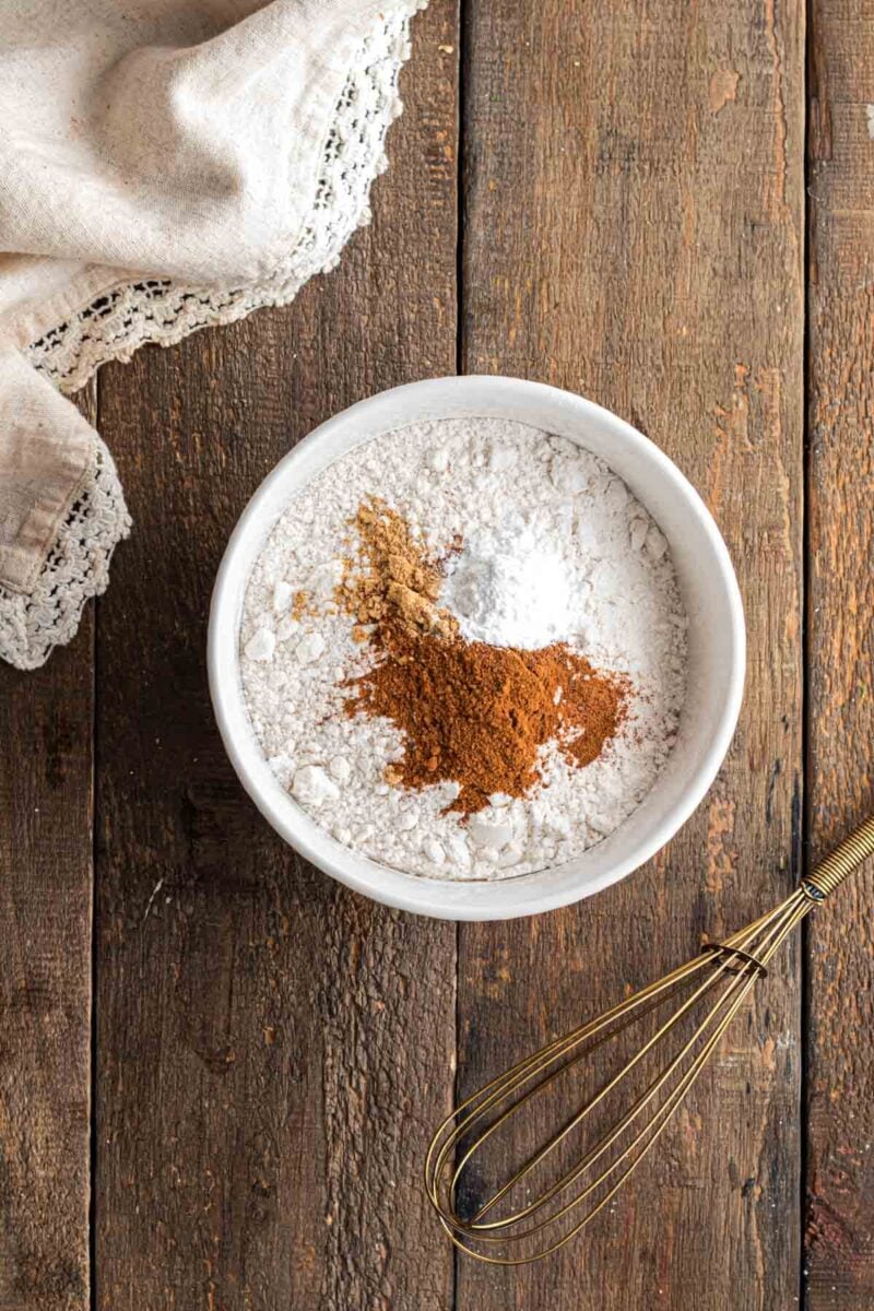 A white bowl with flour, baking powder, cinnamon, and nutmeg on a wooden surface, next to a metal whisk and a cloth napkin with lace trim.