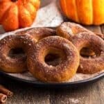 A plate of cinnamon sugar pumpkin donuts on a wooden table with two pumpkins and cinnamon sticks in the background.