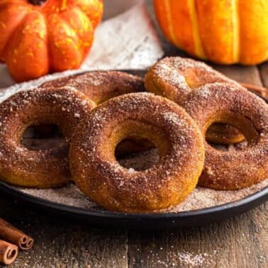 A plate of cinnamon sugar pumpkin donuts on a wooden table with two pumpkins and cinnamon sticks in the background.