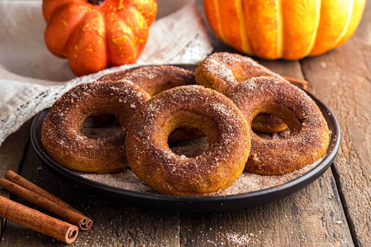 A plate of cinnamon sugar pumpkin donuts on a wooden table with two pumpkins and cinnamon sticks in the background.