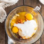 A glass mixing bowl with eggs, flour, milk, and brown sugar on a wooden surface, ready to be mixed; a whisk and bowl of flour nearby.