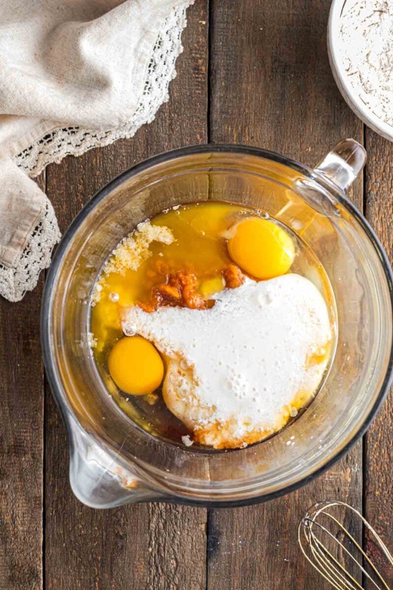 A glass mixing bowl with eggs, flour, milk, and brown sugar on a wooden surface, ready to be mixed; a whisk and bowl of flour nearby.