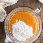 A glass mixing bowl with orange batter and a pile of white flour on top, set on a wooden surface next to a whisk and a cloth.