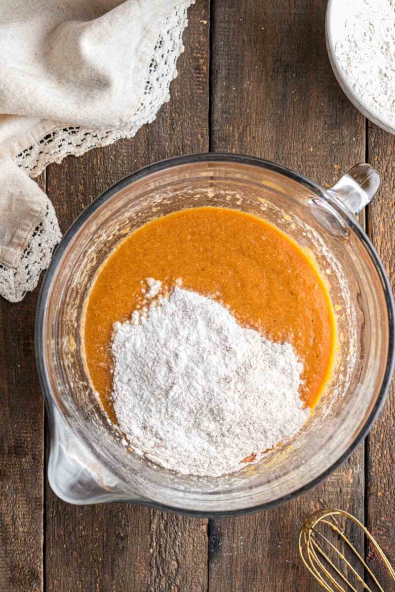 A glass mixing bowl with orange batter and a pile of white flour on top, set on a wooden surface next to a whisk and a cloth.