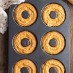 A six-cavity donut pan filled with raw pumpkin batter, with a piping bag nearby on a wooden surface and a lace-edged cloth in the corner.