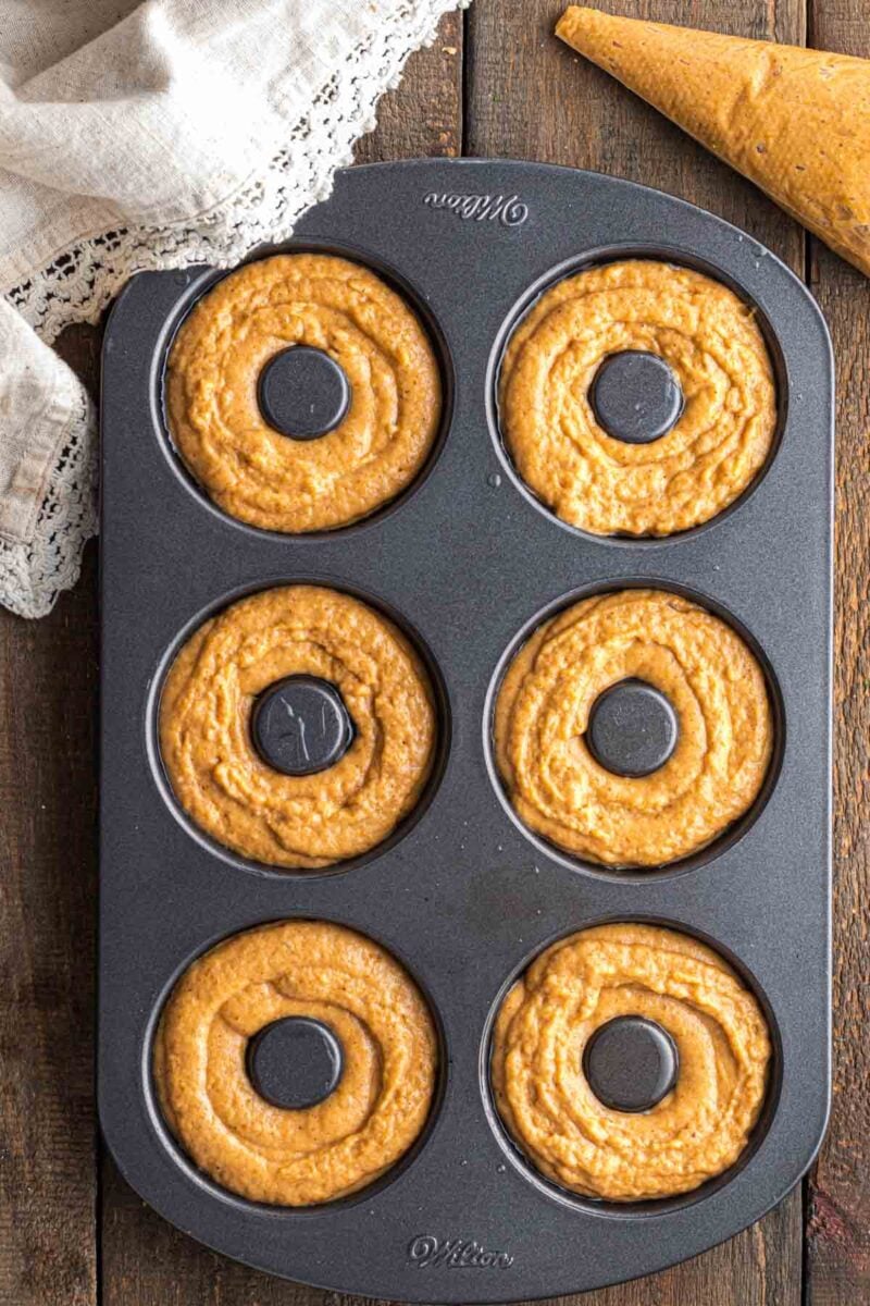 A six-cavity donut pan filled with raw pumpkin batter, with a piping bag nearby on a wooden surface and a lace-edged cloth in the corner.