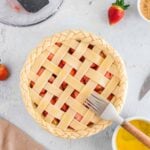 Unbaked strawberry pie with a lattice crust is brushed with egg wash; strawberries, sugar, and baking tools are nearby on a light surface.