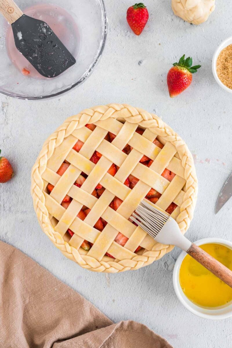 Unbaked strawberry pie with a lattice crust is brushed with egg wash; strawberries, sugar, and baking tools are nearby on a light surface.