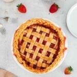 A baked strawberry pie with a braided lattice crust sits on a light surface, surrounded by fresh strawberries, a fork, plates, and a beige napkin.