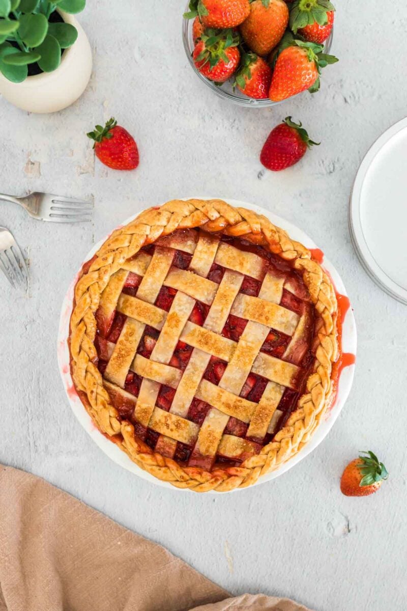 A baked strawberry pie with a braided lattice crust sits on a light surface, surrounded by fresh strawberries, a fork, plates, and a beige napkin.