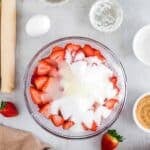 Sliced strawberries in a glass bowl topped with sugar, surrounded by baking ingredients including an egg, measuring cups, pie dough, and a rolling pin on a light surface.