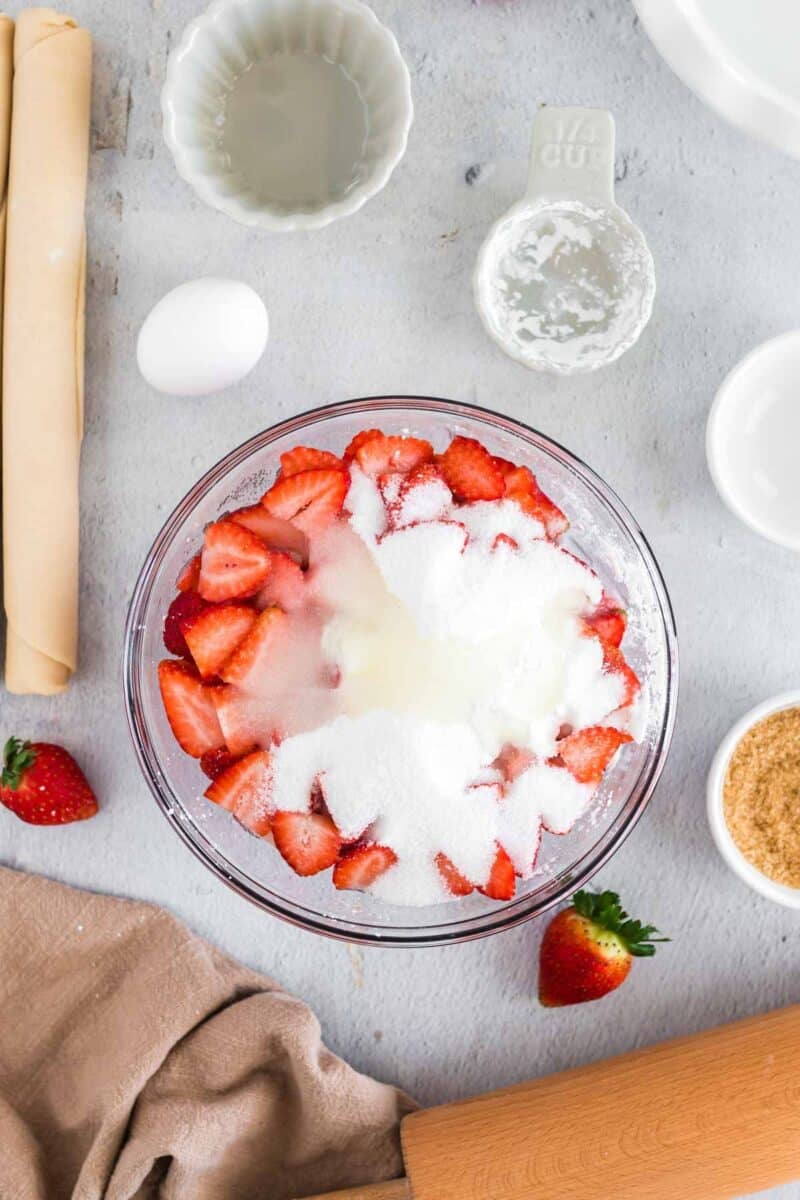 Sliced strawberries in a glass bowl topped with sugar, surrounded by baking ingredients including an egg, measuring cups, pie dough, and a rolling pin on a light surface.
