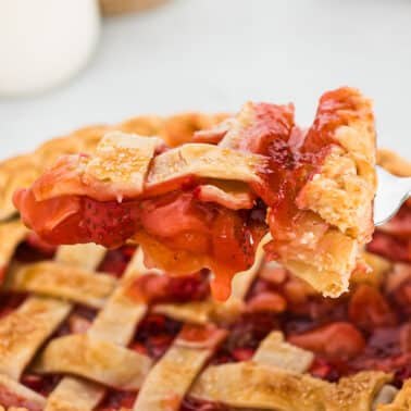 A fork holds a slice of lattice-topped strawberry pie above the remaining pie, with filling dripping from the crust.