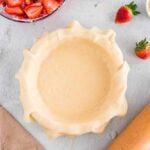 Unbaked pie crust in a dish on a light countertop, surrounded by sliced strawberries, a rolling pin, whole strawberries, and a tan cloth.