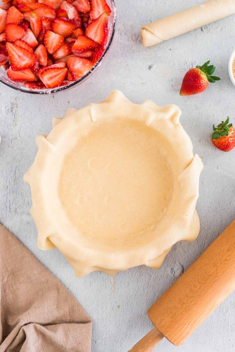 Unbaked pie crust in a dish on a light countertop, surrounded by sliced strawberries, a rolling pin, whole strawberries, and a tan cloth.
