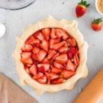 Unbaked strawberry pie with sliced strawberries in a pastry crust, surrounded by a rolling pin, strawberries, sugar, dough, and mixing bowl on a light surface.