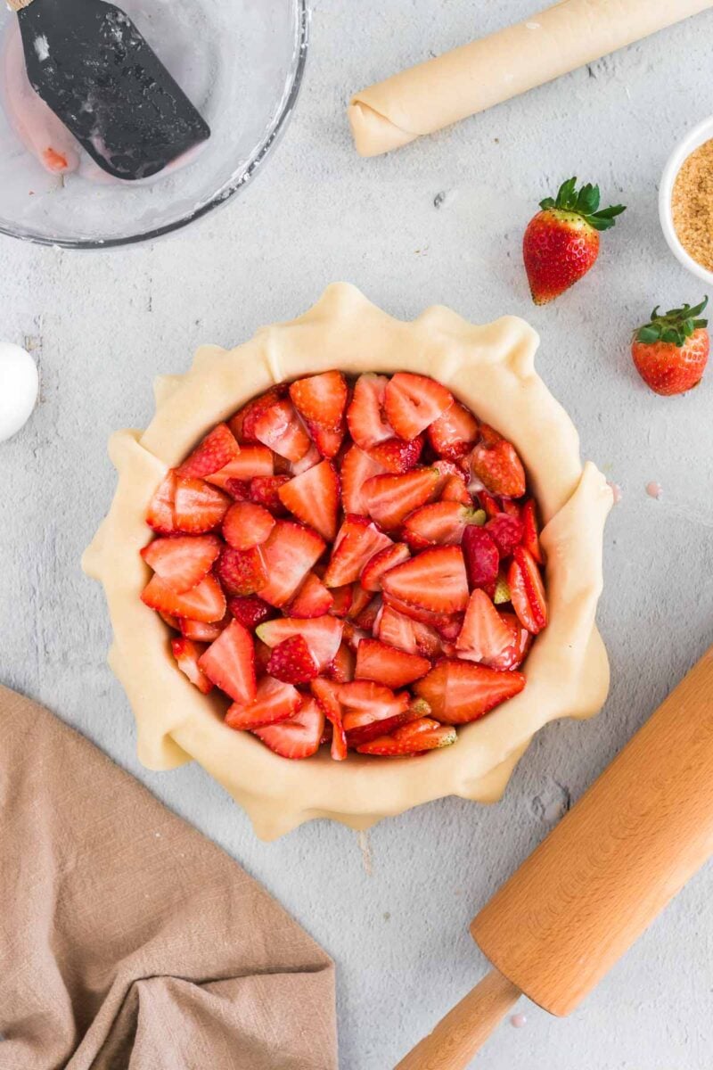 Unbaked strawberry pie with sliced strawberries in a pastry crust, surrounded by a rolling pin, strawberries, sugar, dough, and mixing bowl on a light surface.