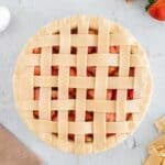 Unbaked strawberry pie with a lattice crust sits on a light surface, surrounded by strawberries, dough strips, a brown cloth, and an egg.
