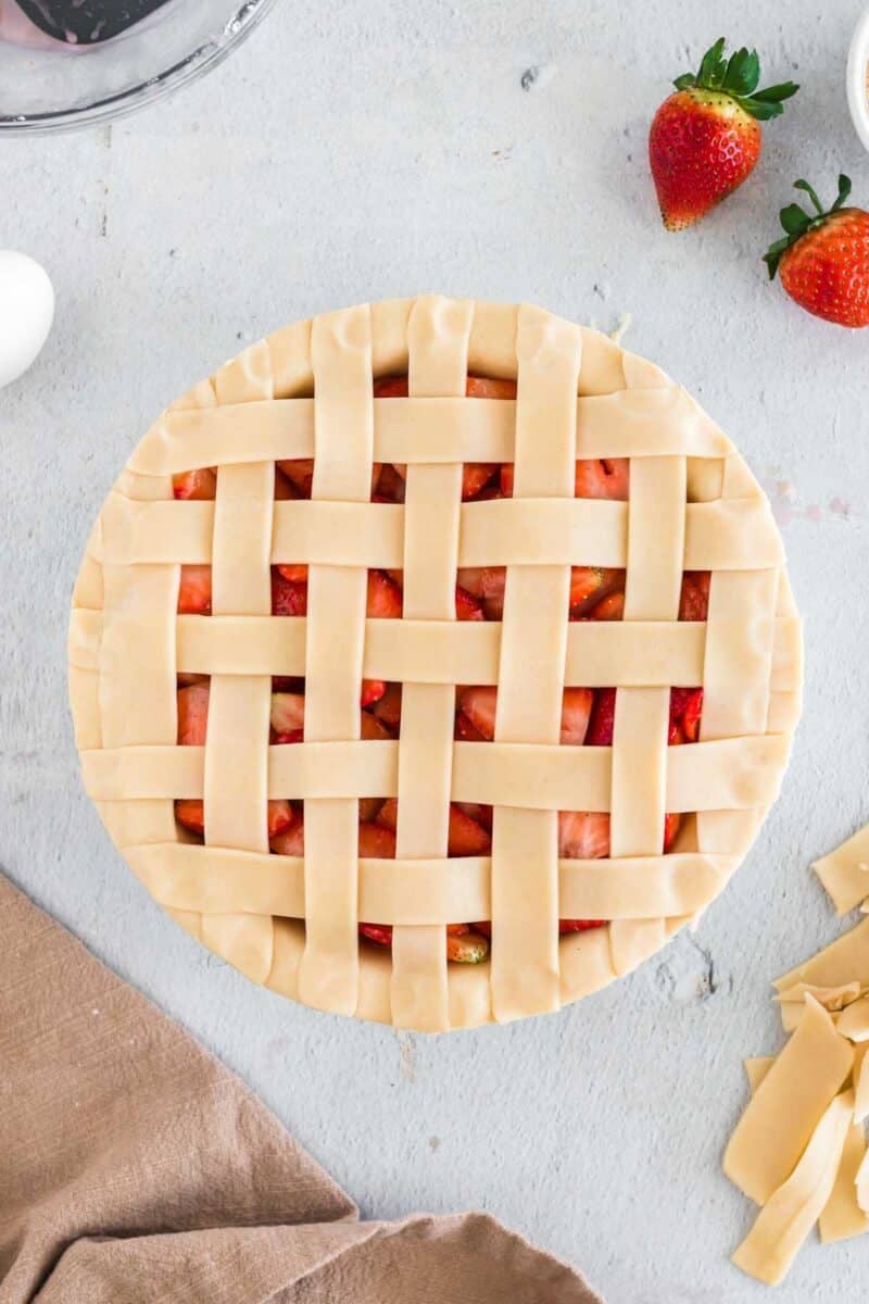 Unbaked strawberry pie with a lattice crust sits on a light surface, surrounded by strawberries, dough strips, a brown cloth, and an egg.