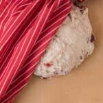 A ball of bread dough with dried fruit rests on parchment paper, partially covered by a red and white striped cloth.