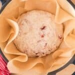 Round loaf of unbaked bread dough with cranberries and nuts sits on parchment paper in a black Dutch oven, with a red-striped cloth nearby.