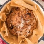 Round loaf of rustic bread with a golden crust sits on parchment paper in a large pot, with red striped towel and small bowls of ingredients nearby.