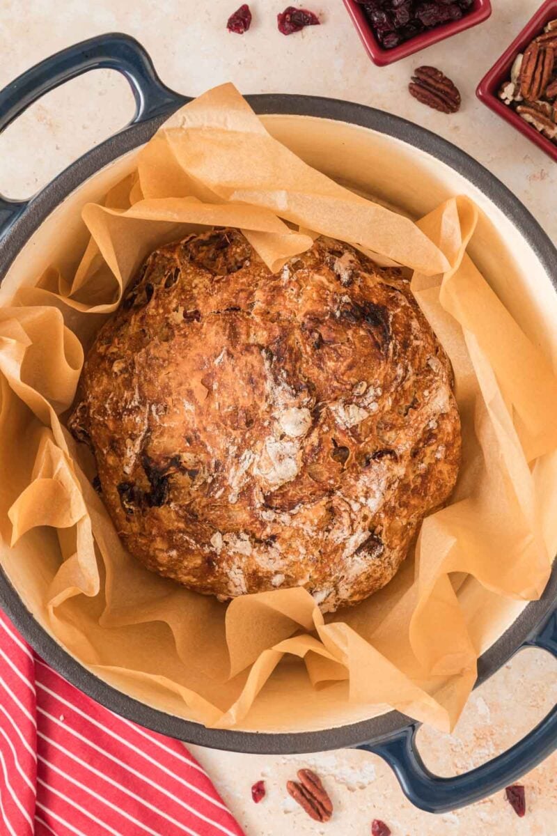 Round loaf of rustic bread with a golden crust sits on parchment paper in a large pot, with red striped towel and small bowls of ingredients nearby.