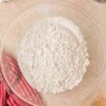 A glass bowl filled with flour sits on a countertop next to a red striped towel, wooden spoon, and small bowls of ingredients.