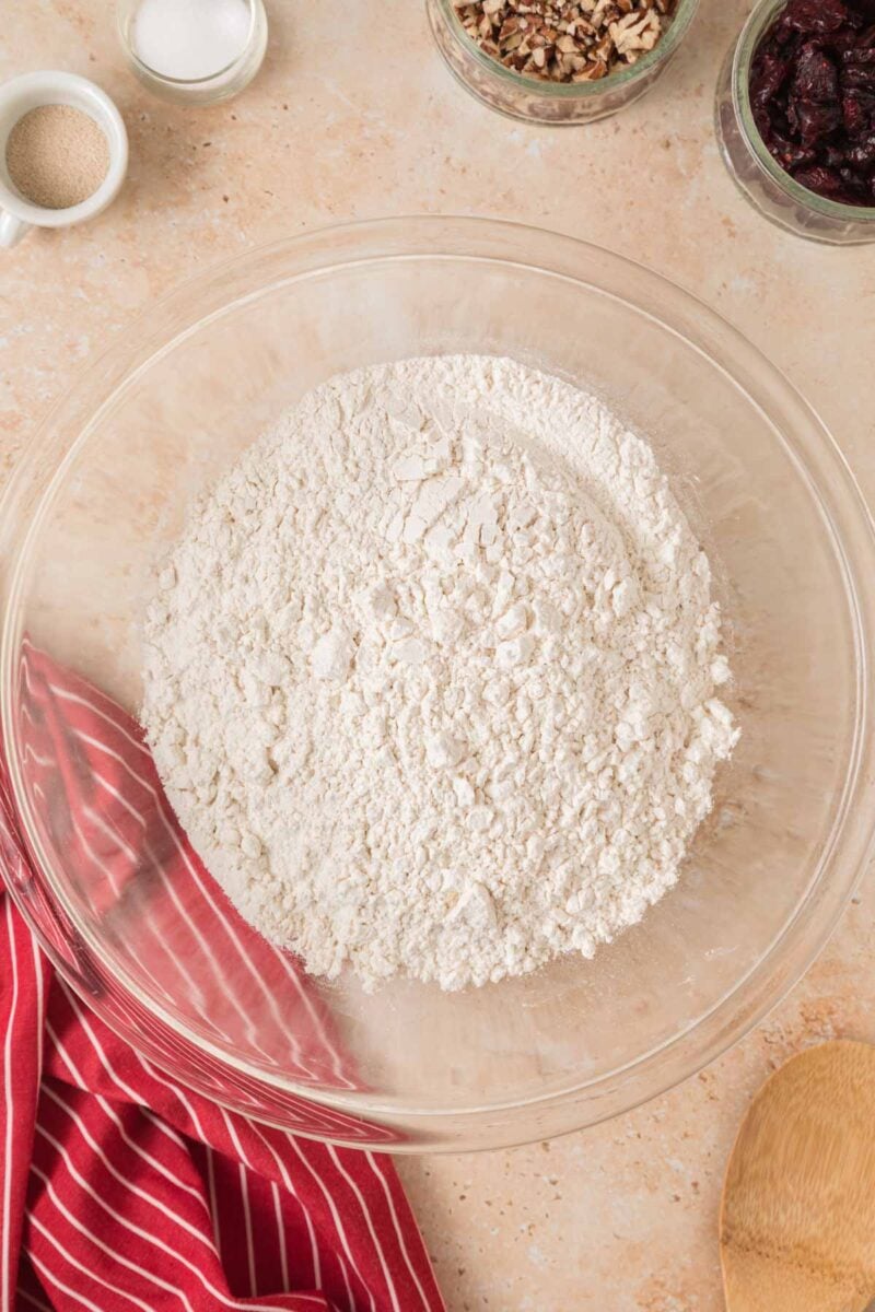 A glass bowl filled with flour sits on a countertop next to a red striped towel, wooden spoon, and small bowls of ingredients.