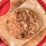 A round loaf of rustic bread with a golden crust sits on parchment paper atop a wooden cutting board, near a red cloth and a small bowl of dried cranberries.