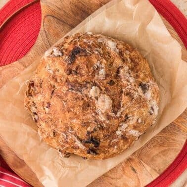 A round loaf of rustic bread with a golden crust sits on parchment paper atop a wooden cutting board, near a red cloth and a small bowl of dried cranberries.