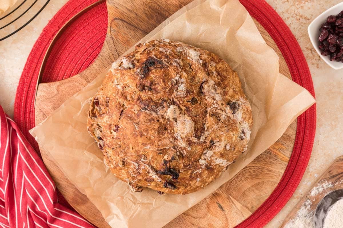 A round loaf of rustic bread with a golden crust sits on parchment paper atop a wooden cutting board, near a red cloth and a small bowl of dried cranberries.