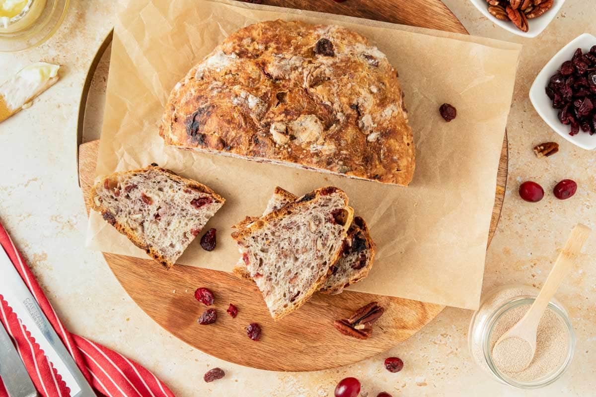 A loaf of rustic bread with cranberries and pecans sits on parchment paper; two slices are cut. Surrounding items include pecans, cranberries, a jar of yeast, and a red-striped towel.