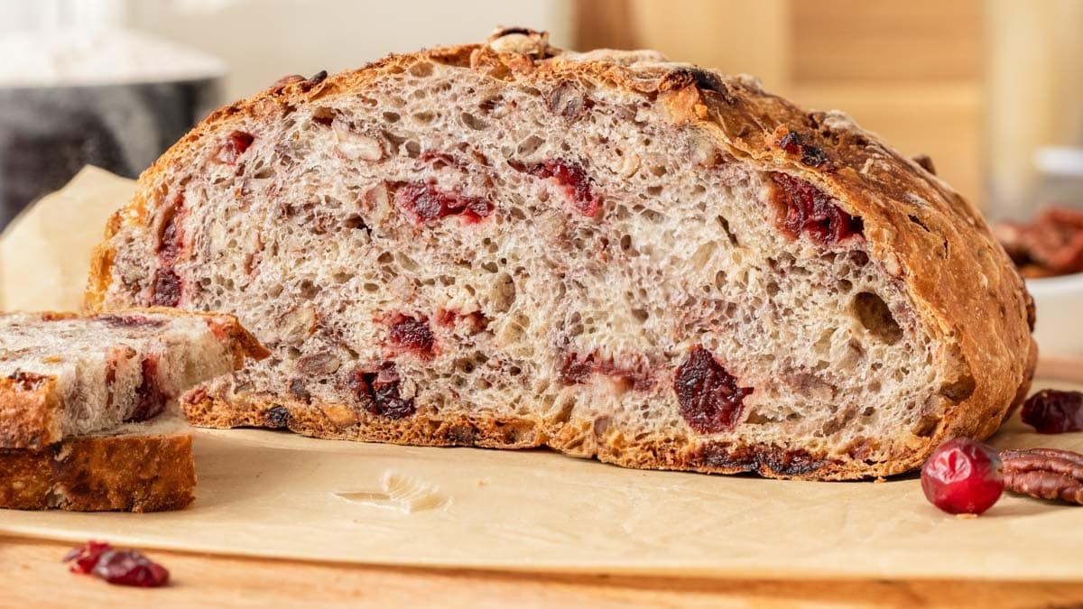 A loaf of rustic bread with visible cranberries and nuts, sliced to show the textured interior, sits on parchment paper on a wooden surface.