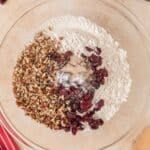 A glass bowl filled with flour, chopped pecans, dried cranberries, yeast, and salt on a light countertop.