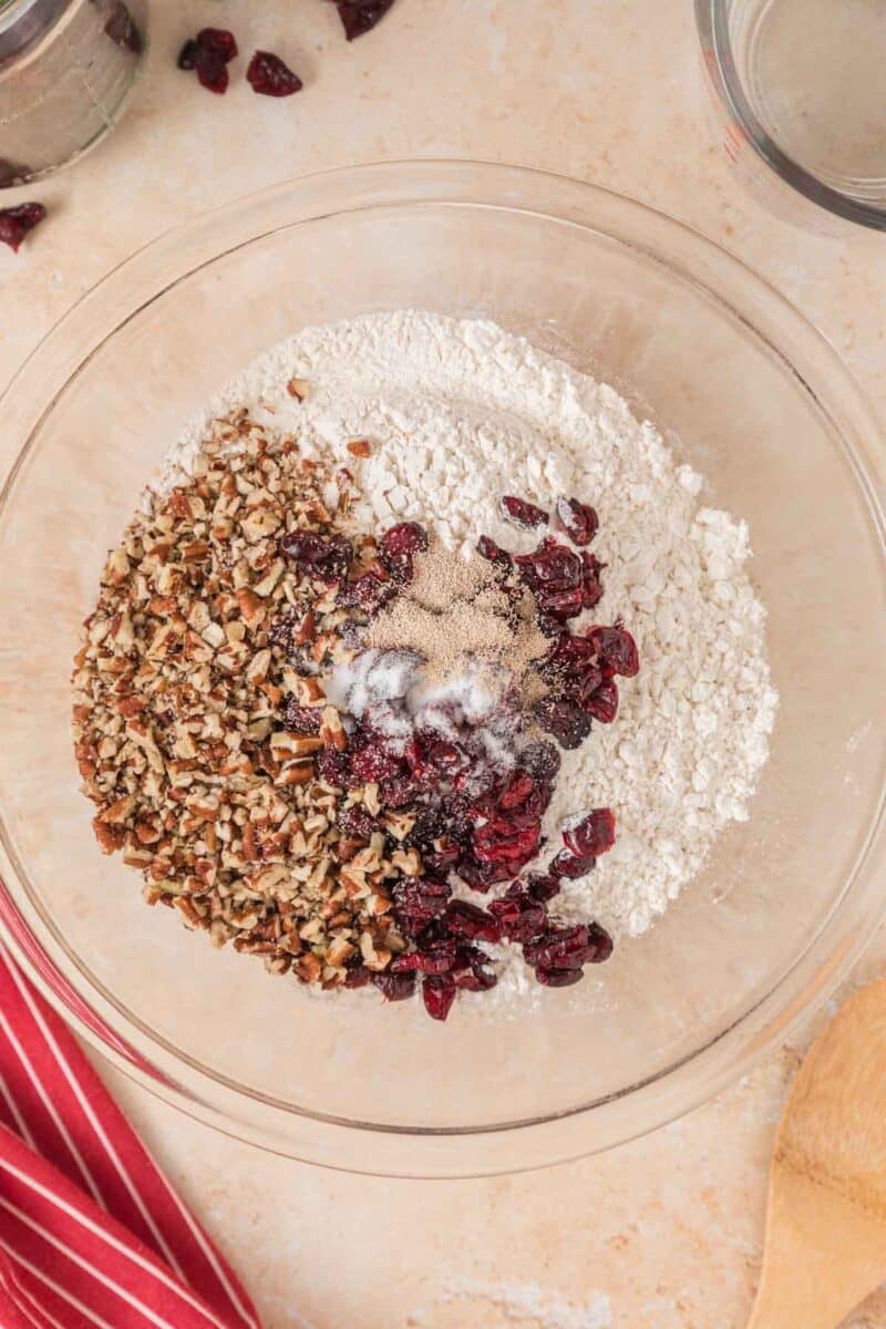 A glass bowl filled with flour, chopped pecans, dried cranberries, yeast, and salt on a light countertop.