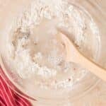 A glass bowl with flour, chopped nuts, and water being mixed with a wooden spoon on a light countertop next to a red striped cloth.