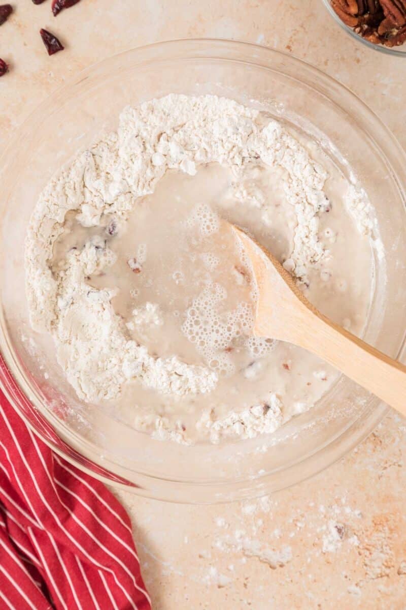 A glass bowl with flour, chopped nuts, and water being mixed with a wooden spoon on a light countertop next to a red striped cloth.
