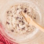 A glass bowl of mixed dough with dried fruit and nuts, a wooden spoon inside, and a red striped cloth nearby on a light surface.