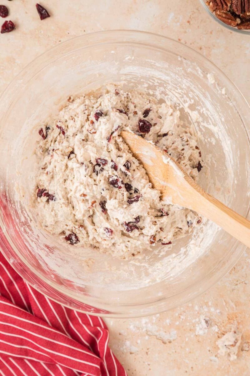 A glass bowl of mixed dough with dried fruit and nuts, a wooden spoon inside, and a red striped cloth nearby on a light surface.
